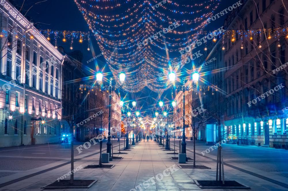На фотографии изображена одна из центральных улиц Санкт-Петербурга, украшенная праздничными гирляндами. Светящиеся огни висят над тротуаром, создавая атмосферу новогоднего или рождественского праздника. Улица освещена фонарями, а здания по бокам украшены подсветкой, подчеркивающей архитектурные детали. Праздничное убранство города придает этому вечернему пейзажу волшебный вид.
